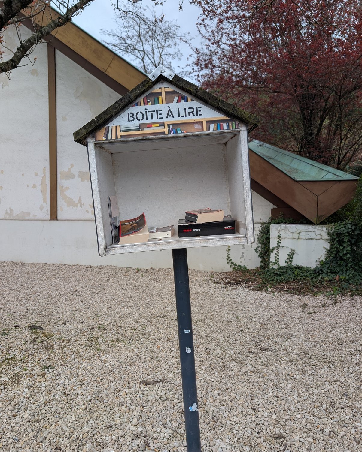 A small outdoor book exchange box shaped like a little house, with a peaked roof and the French label “BOÎTE À LIRE” across the top. It stands on a black post over a gravel area beside a building, with a few books visible inside and trees in the background.