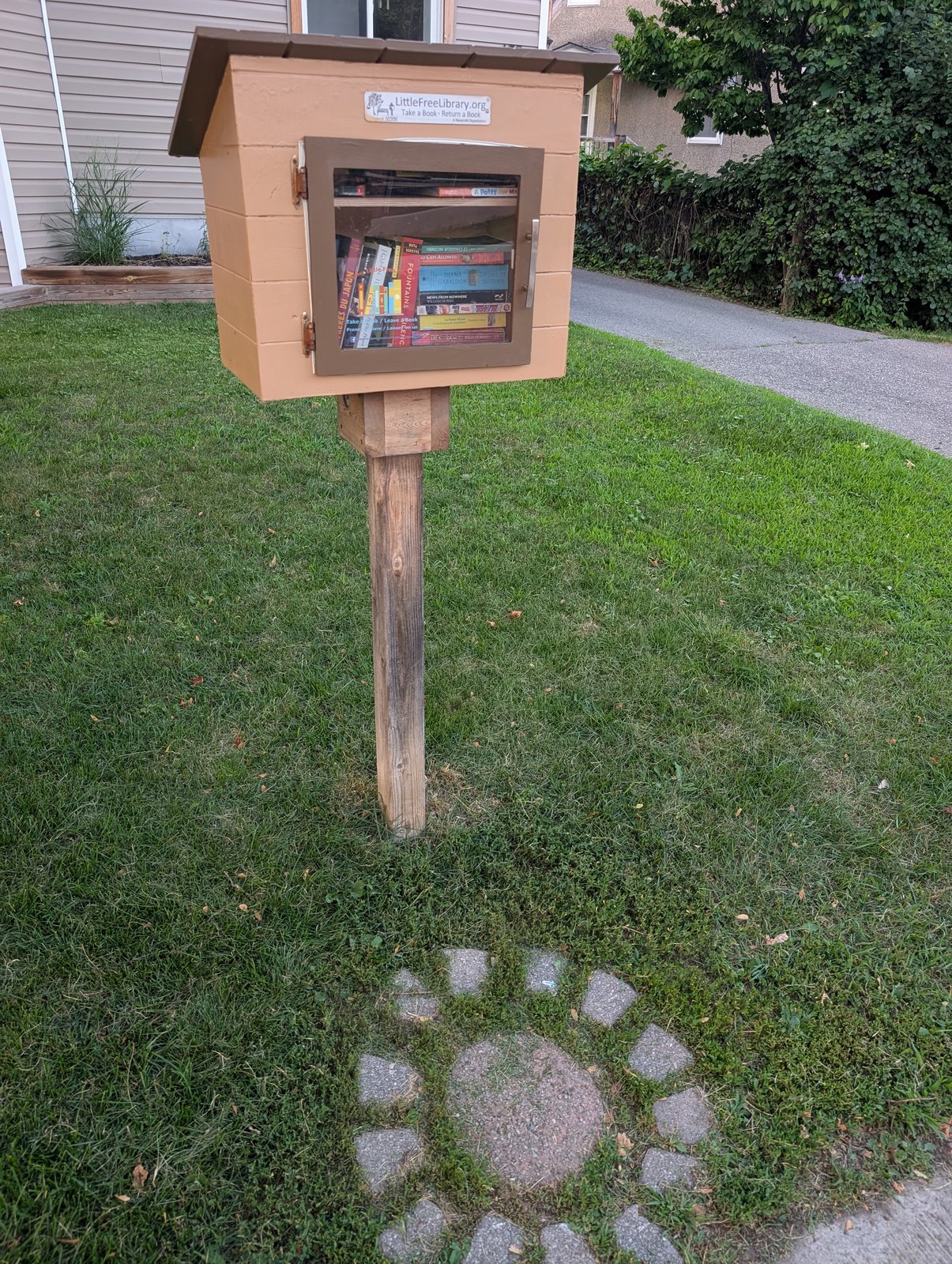 A peach-colored Little Free Library box with a dark brown roof and glass-front door stands on a wooden post in a front lawn. It sits beside a sidewalk near a house and hedges, with books visible inside and a small LittleFreeLibrary.org sign on the front.