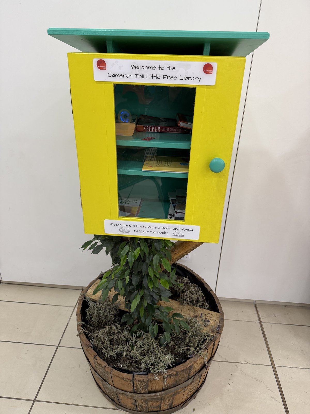 A bright yellow little free library box with a teal roof and door, mounted above a planter base in a barrel. It stands indoors against a plain light wall, with books visible through the front window and a small sign inviting visitors to take and leave books.