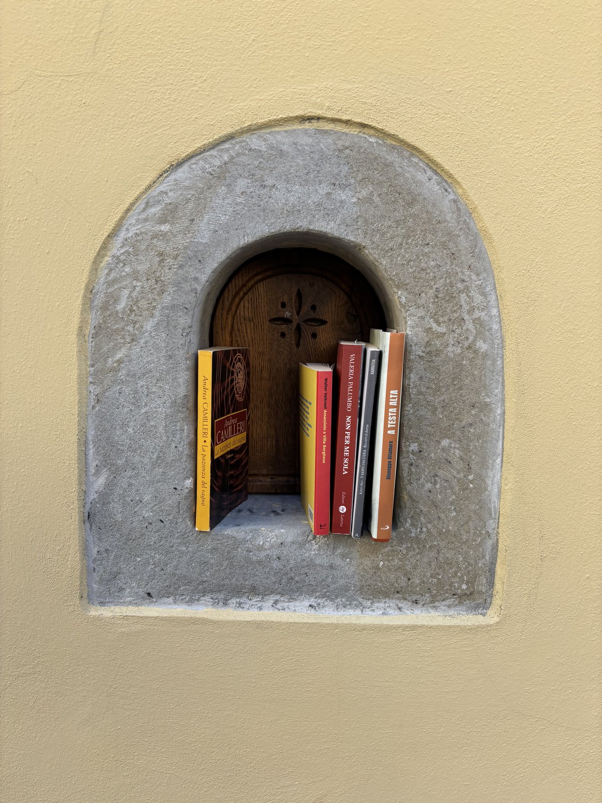 A small free-book exchange is built into a rounded stone-framed niche in a pale yellow wall. Several paperback books stand upright inside the recessed wooden compartment, giving it the feel of a tiny neighborhood library.
