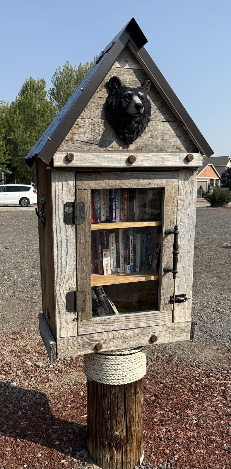 A rustic wooden book exchange box with a steep metal roof and a decorative black bear head mounted on the front. It stands outdoors on a wooden post wrapped with rope, with glass-fronted shelves holding books inside.