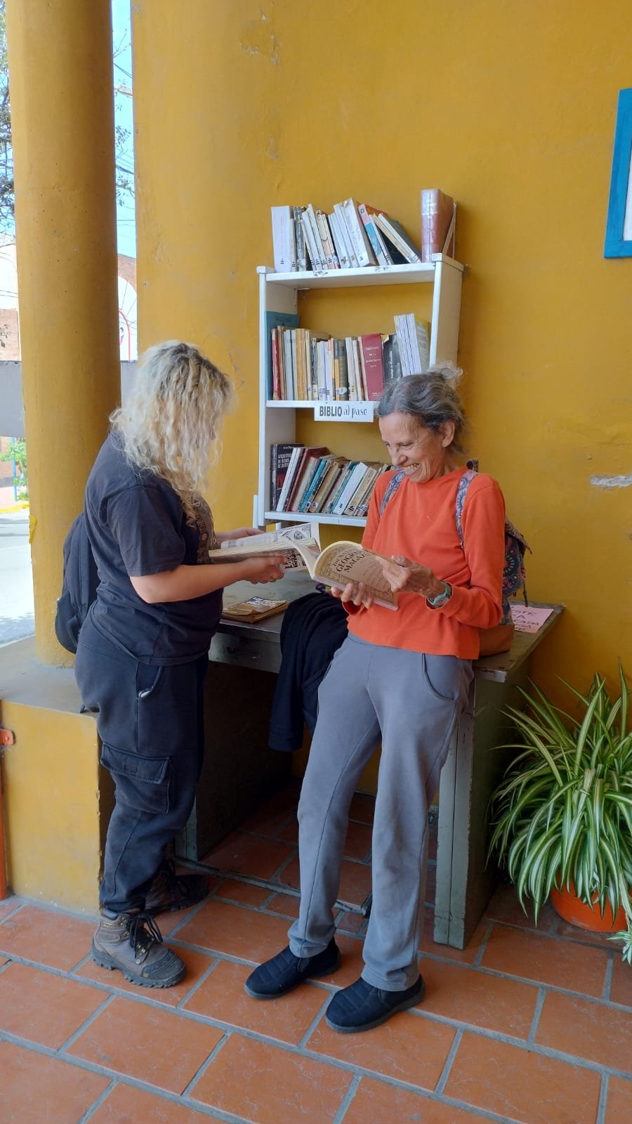 A small community book exchange shelf built against a bright yellow wall, with several rows of books and a handwritten sign. Two people stand at the shelf reading and browsing books beside a potted plant and a tiled porch.