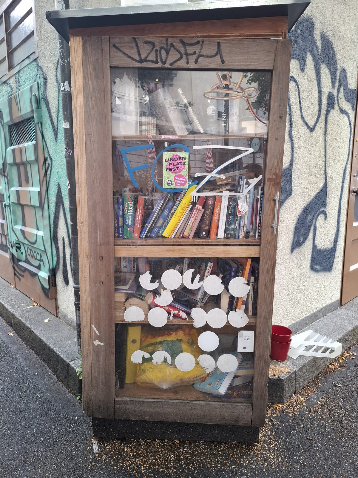 A tall wooden, glass-fronted book exchange cabinet stands against a graffiti-covered urban wall at Hallwylplatz in Zurich. It is densely packed with books in several languages, with colorful posters, stickers, and painted markings covering the doors and surrounding facade.