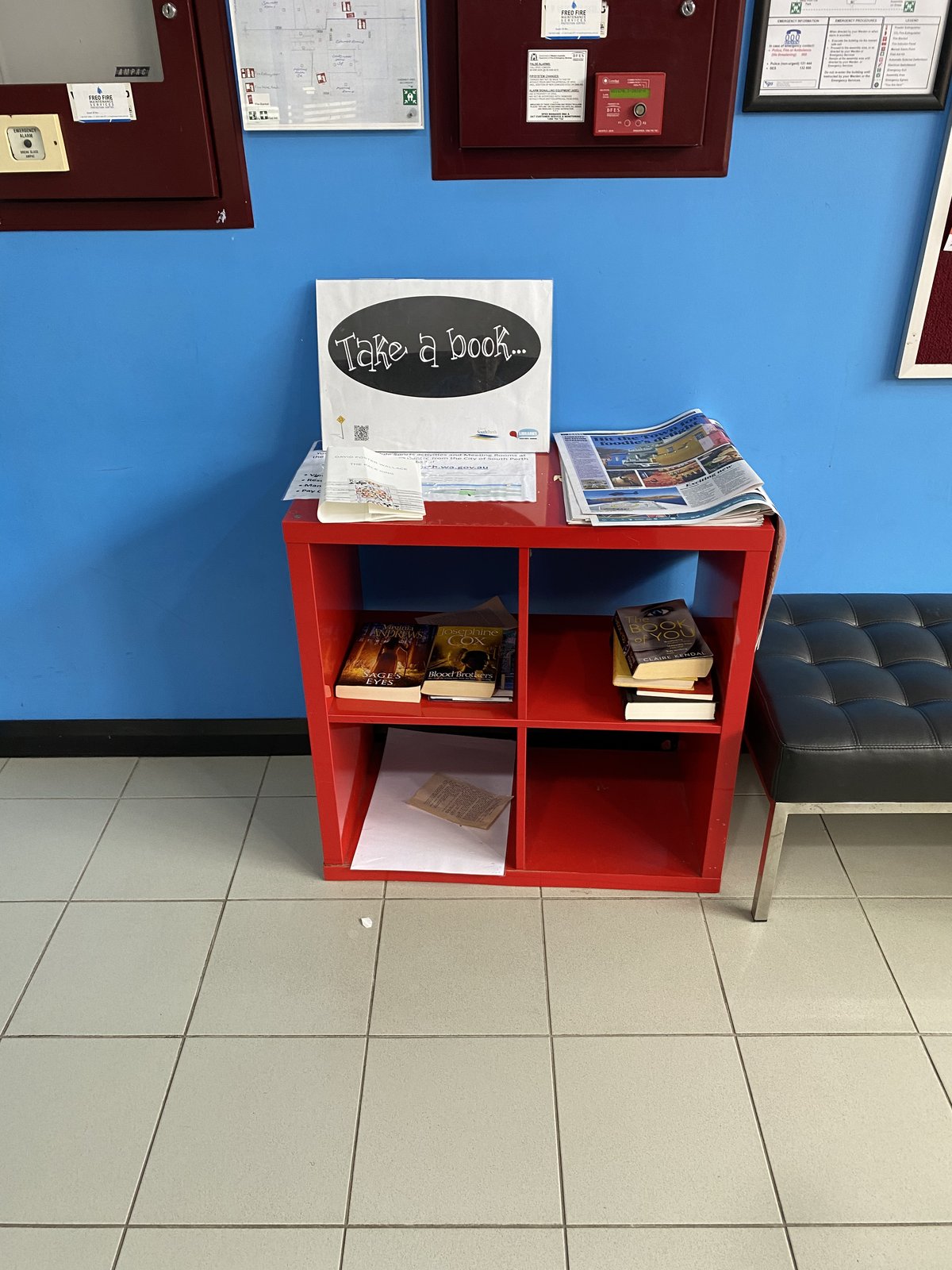 A bright red cubby bookshelf sits against a blue indoor wall at Manning Road, offering a small selection of donated books and newspapers. A prominent “Take a book...” sign and nearby seating make it feel like a welcoming reading exchange in a community waiting area.