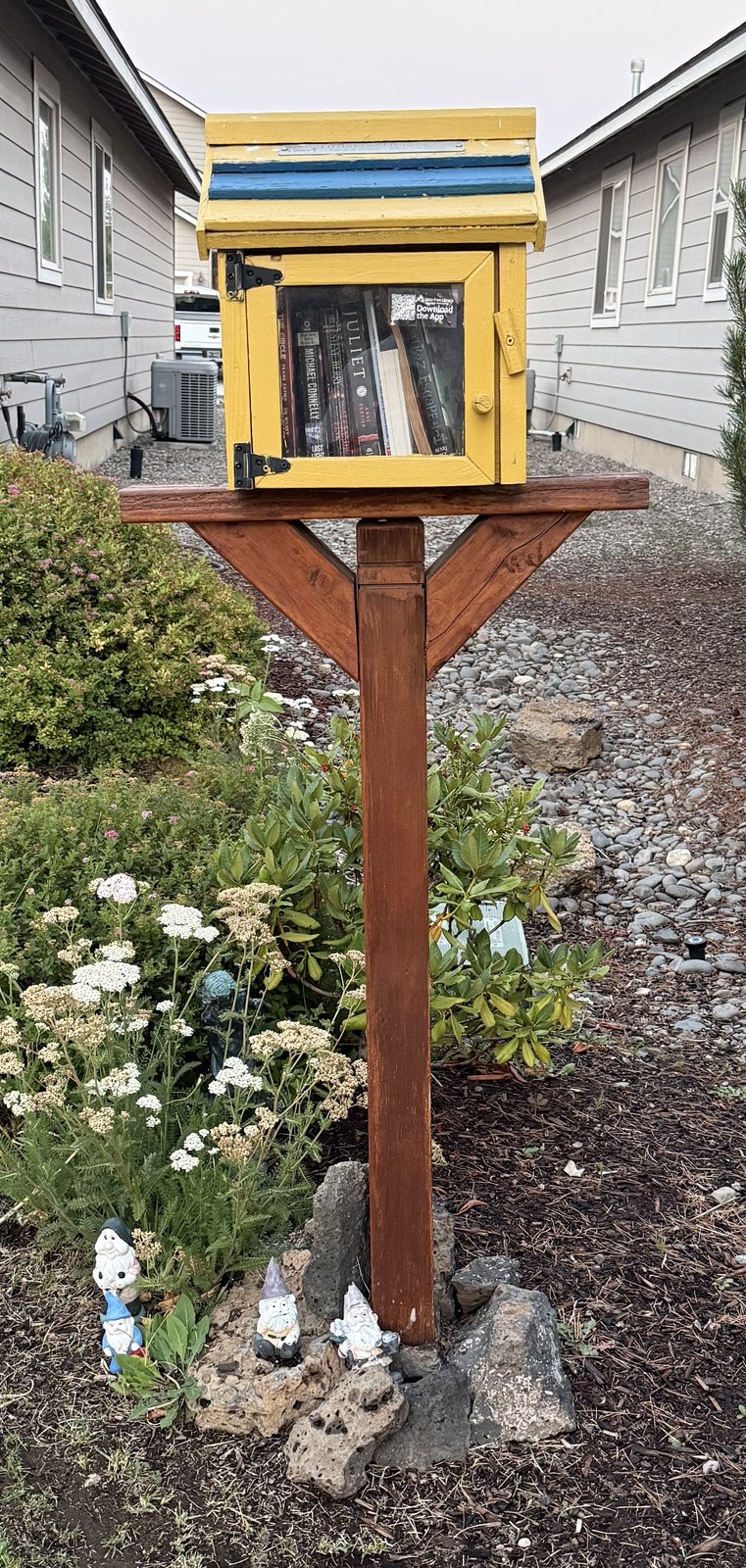 A bright yellow community book exchange with blue trim and a small glass-front door, mounted on a sturdy wooden post. It stands beside a flower garden and rock landscaping, with several garden gnomes gathered at its base.