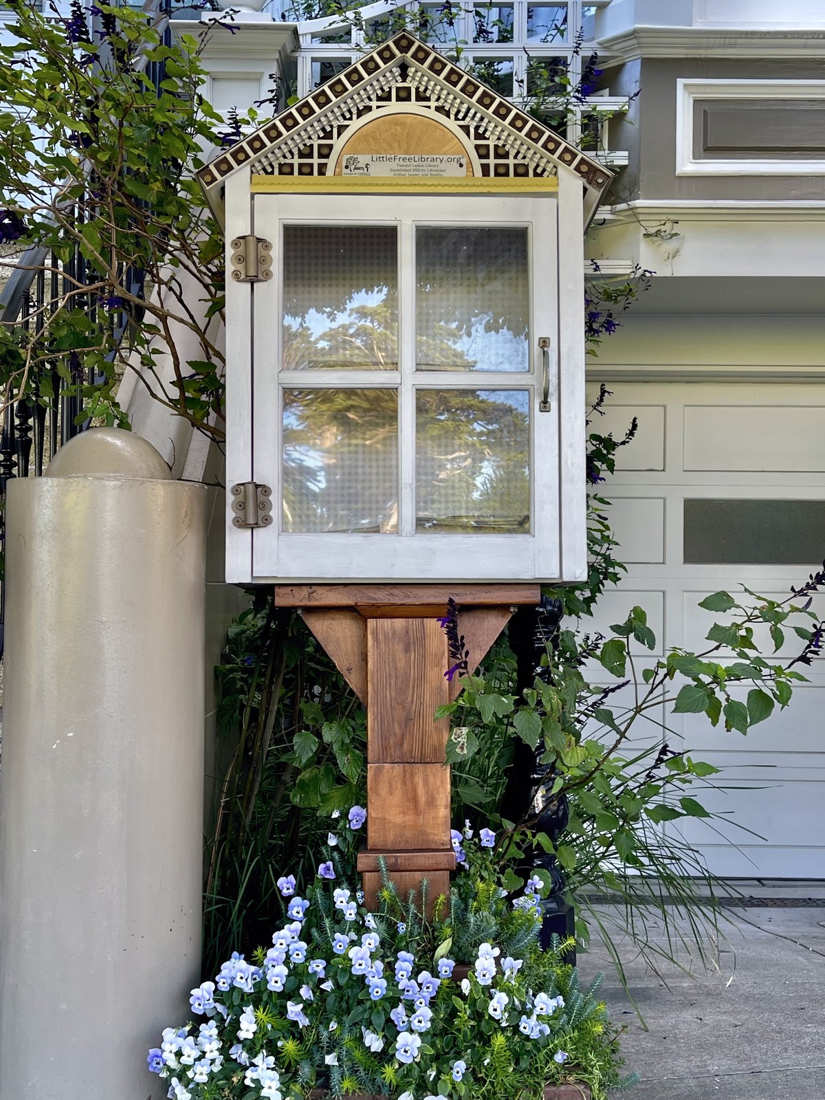 A white, house-shaped Little Free Library features a decorative gabled roof, patterned trim, and glass-paneled double doors. It stands on a wooden post beside a San Francisco home, framed by climbing vines and a lush planter of purple and white flowers.