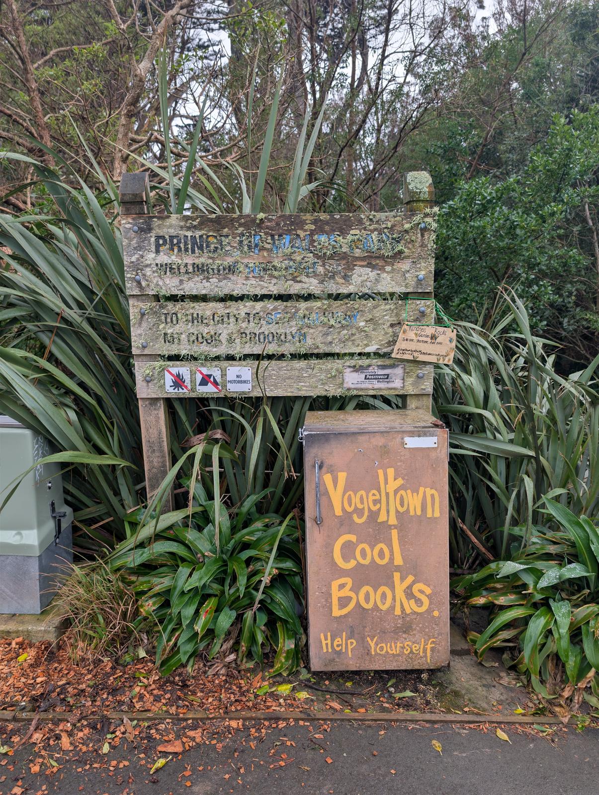 A weathered community book exchange stands beside a path amid dense native greenery. Its rustic wooden sign reads “Yogeltown Cool Books” and “Help Yourself,” with a small metal book box and additional hand-painted notices below.