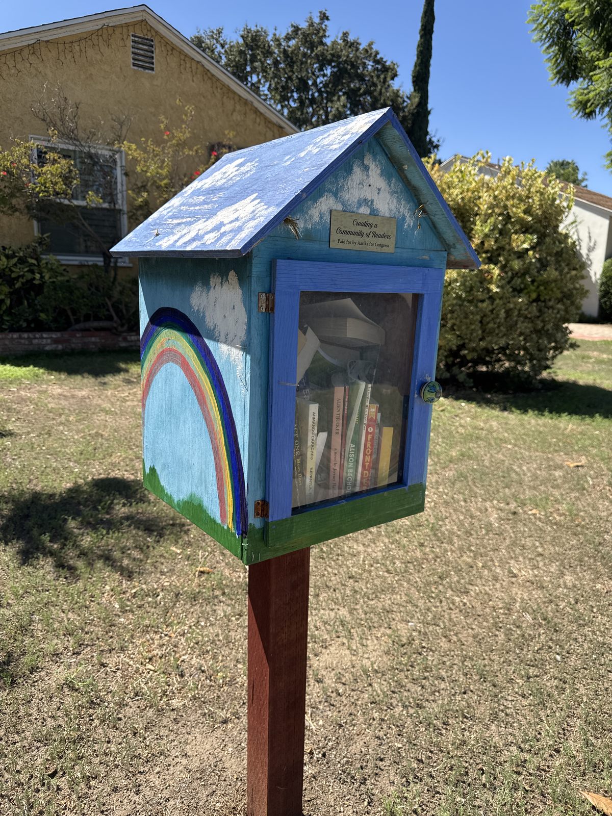 A small, weathered blue wooden book exchange stands on a post in a sunny residential yard. Its glass-fronted cabinet displays books, while a colorful rainbow mural decorates the side and a sign reads “Creating a Community of Readers.”