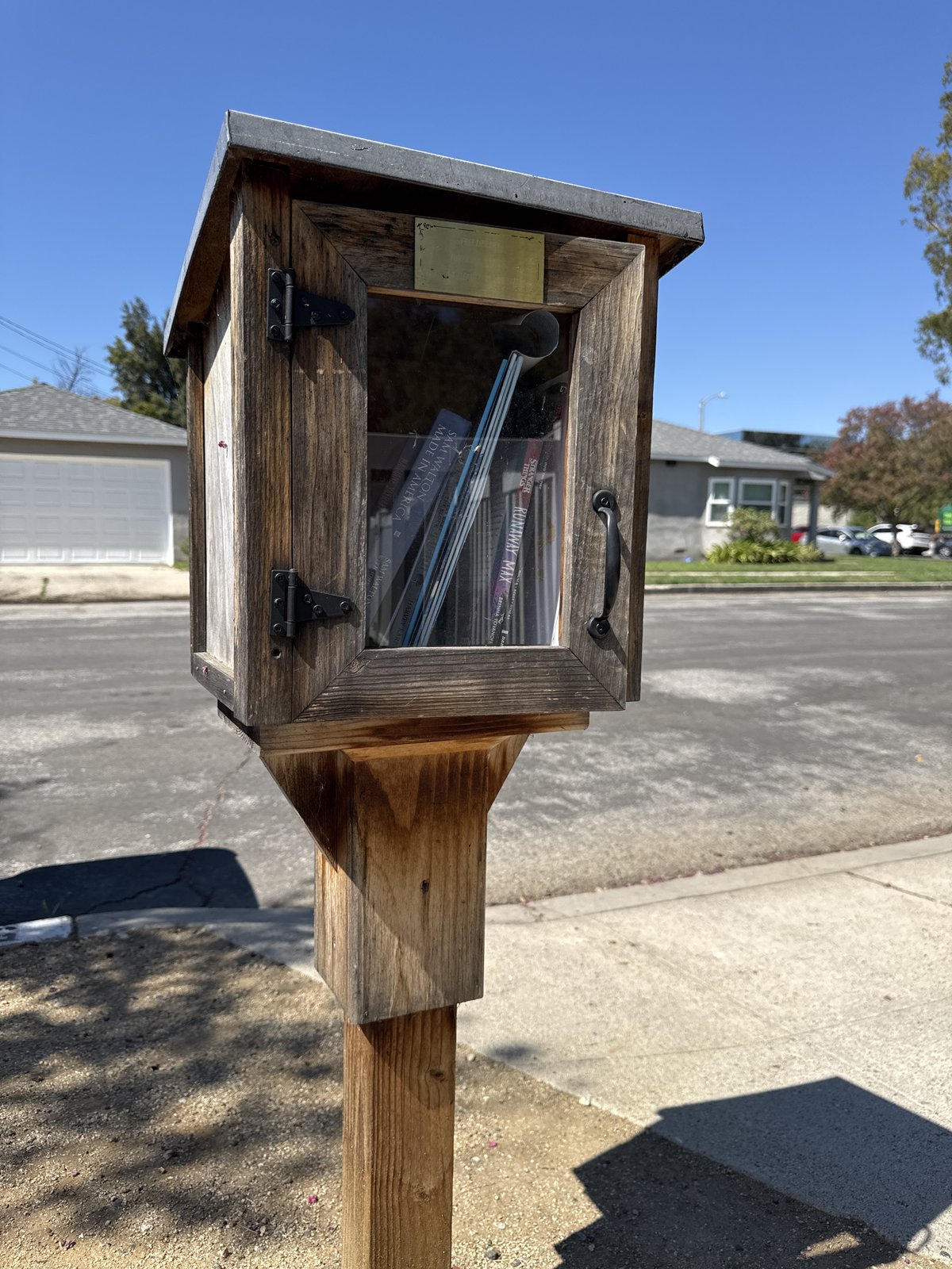 A weathered wooden book exchange box stands on a sturdy post beside a residential sidewalk. Its hinged glass-front door displays a small selection of books, with a broad sloped roof and dark metal hardware adding rustic character.