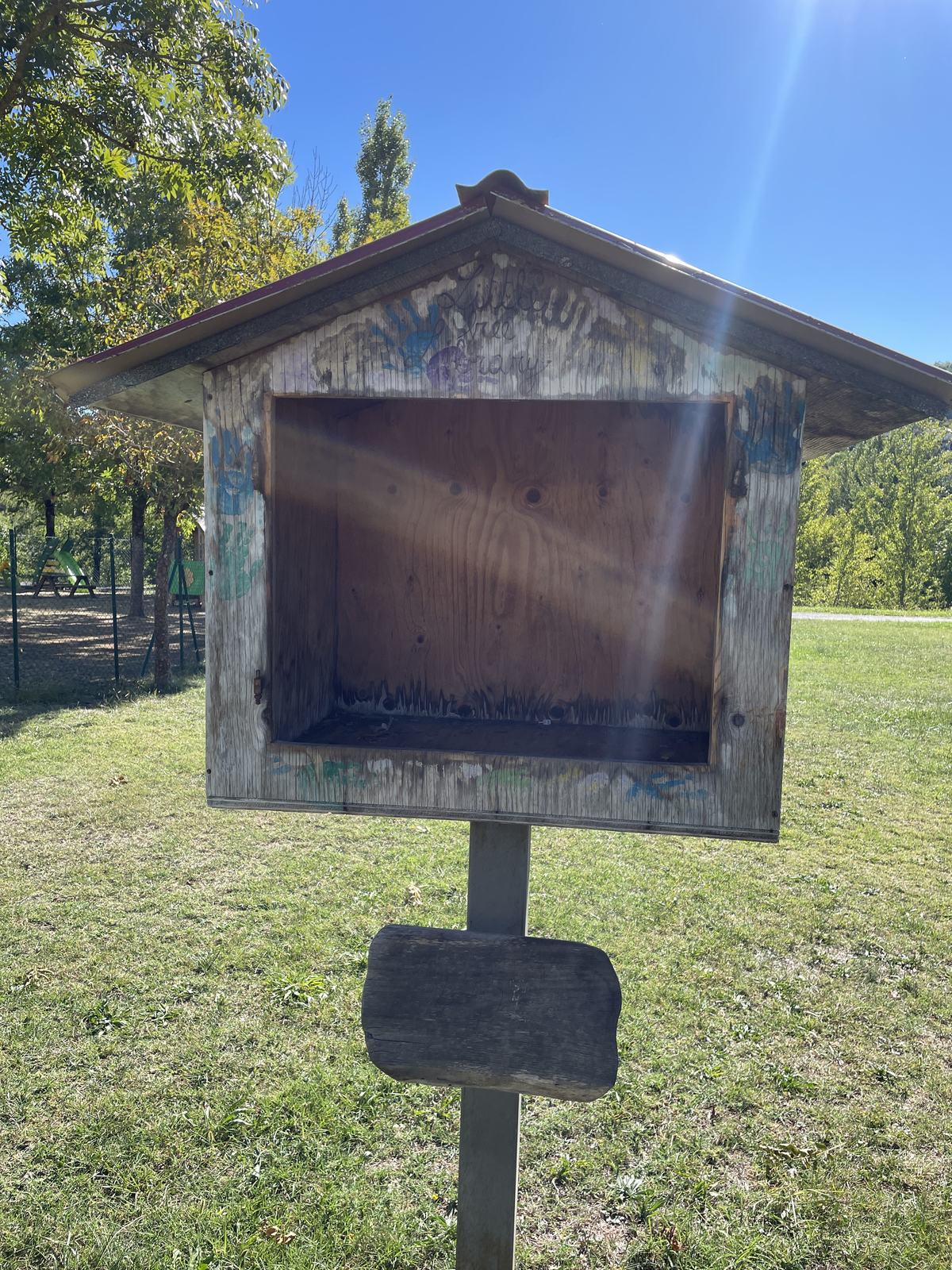 A small, weathered wooden book exchange with a pitched roof and open shelf, mounted on a post in a grassy park. Its unpainted wood is marked with colorful graffiti, and trees, fencing, and playground equipment surround it.
