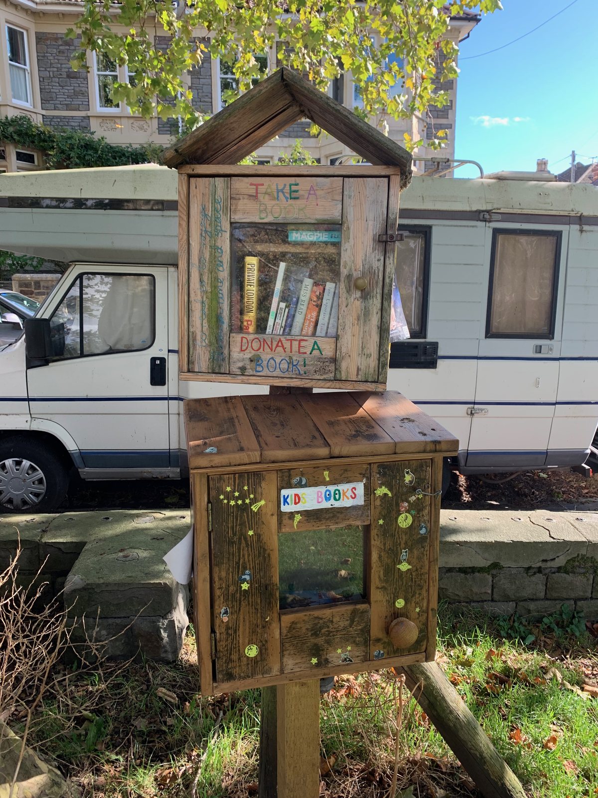 A handmade, weathered wooden book exchange with a peaked roof, glass-fronted shelves, colorful hand-painted “Take a Book” and “Donate a Book” signs, and a lower section labeled “Kids Books.” It stands beside a sidewalk and stone wall in front of a parked camper and a residential home.