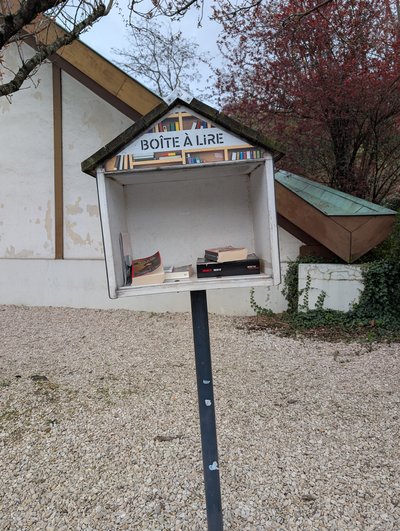 A small outdoor book exchange box shaped like a little house, with a peaked roof and the French label “BOÎTE À LIRE” across the top. It stands on a black post over a gravel area beside a building, with a few books visible inside and trees in the background.