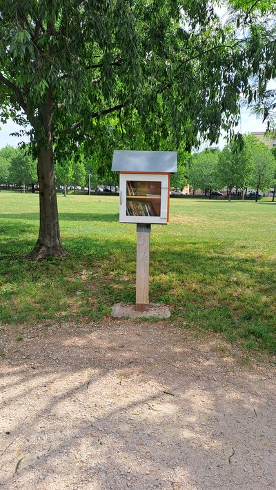 A small freestanding community book exchange box stands on a wooden post in a grassy park under a leafy tree. It has a simple white-and-orange cabinet with a glass front showing books inside and a sloped gray roof.