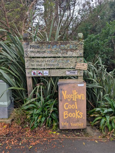 A weathered community book exchange stands beside a path amid dense native greenery. Its rustic wooden sign reads “Yogeltown Cool Books” and “Help Yourself,” with a small metal book box and additional hand-painted notices below.