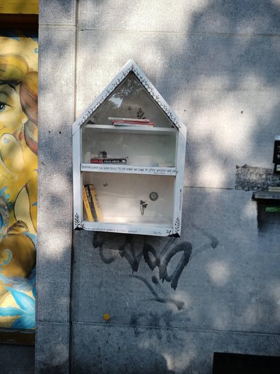 A small white, house-shaped book exchange box is mounted on a gray concrete wall beside a colorful mural. Its glass-fronted shelves hold a few books, with floral decorations and handwritten literary quotes in multiple languages along the trim.