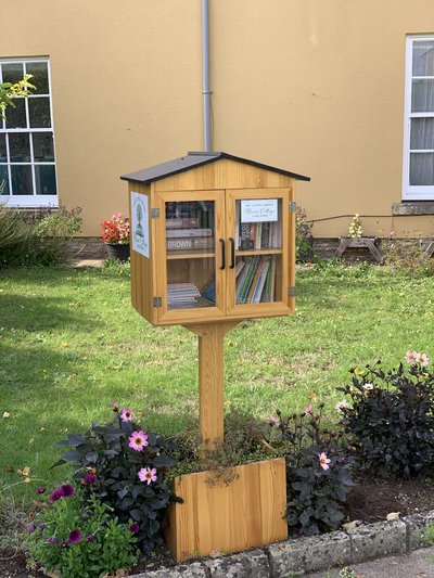 A handsome wooden book exchange cabinet with a pitched roof, glass double doors, and black handles, standing on a post in a flower-filled garden. It is set beside a warm ochre building and stocked with books for adults and children.