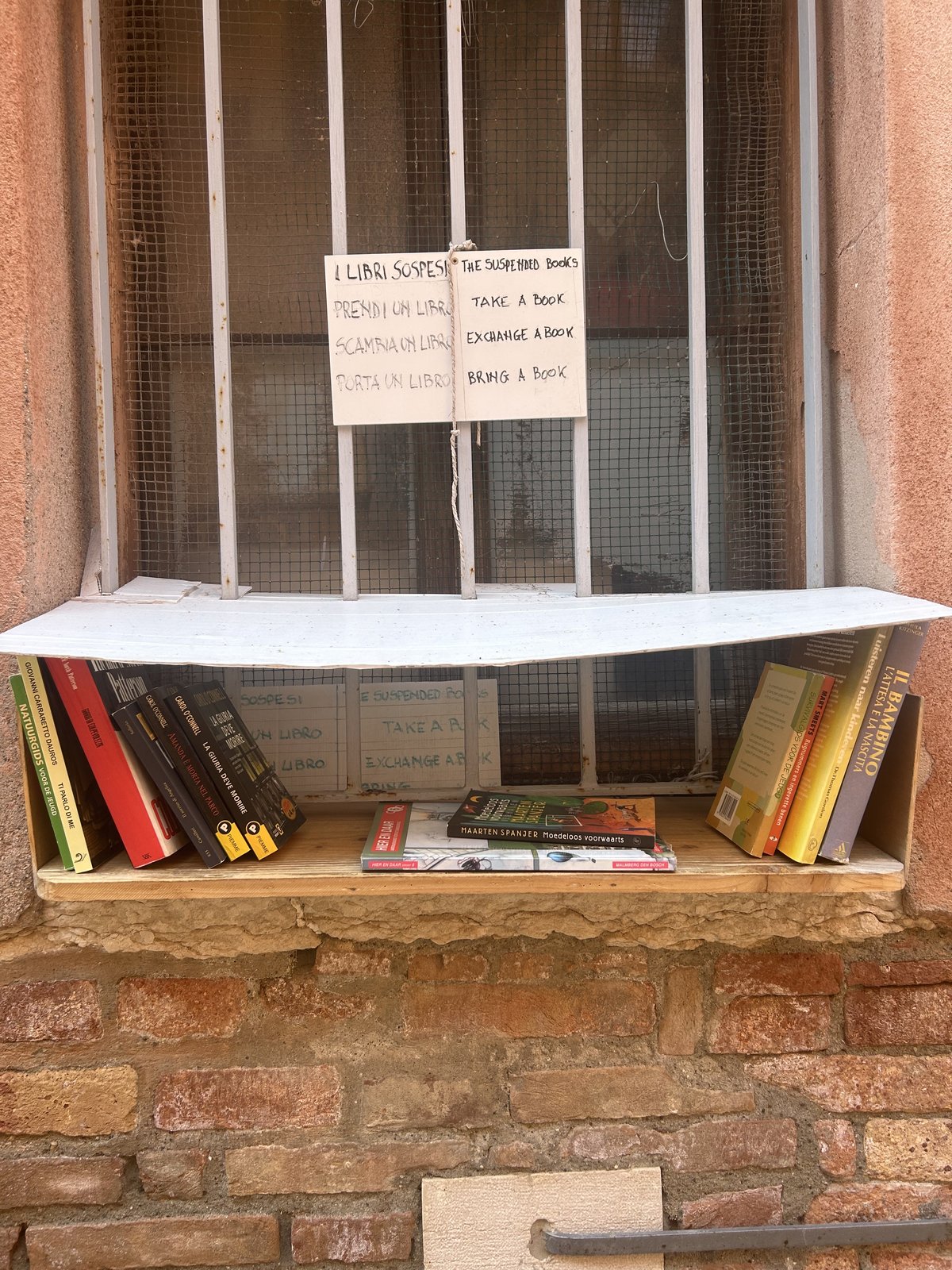 A small free book exchange is built into a barred window set in a weathered brick wall in Venice. A handwritten bilingual sign says “The Suspended Books” and invites visitors to take, exchange, or bring a book, with several paperbacks lined up on the shelf below.